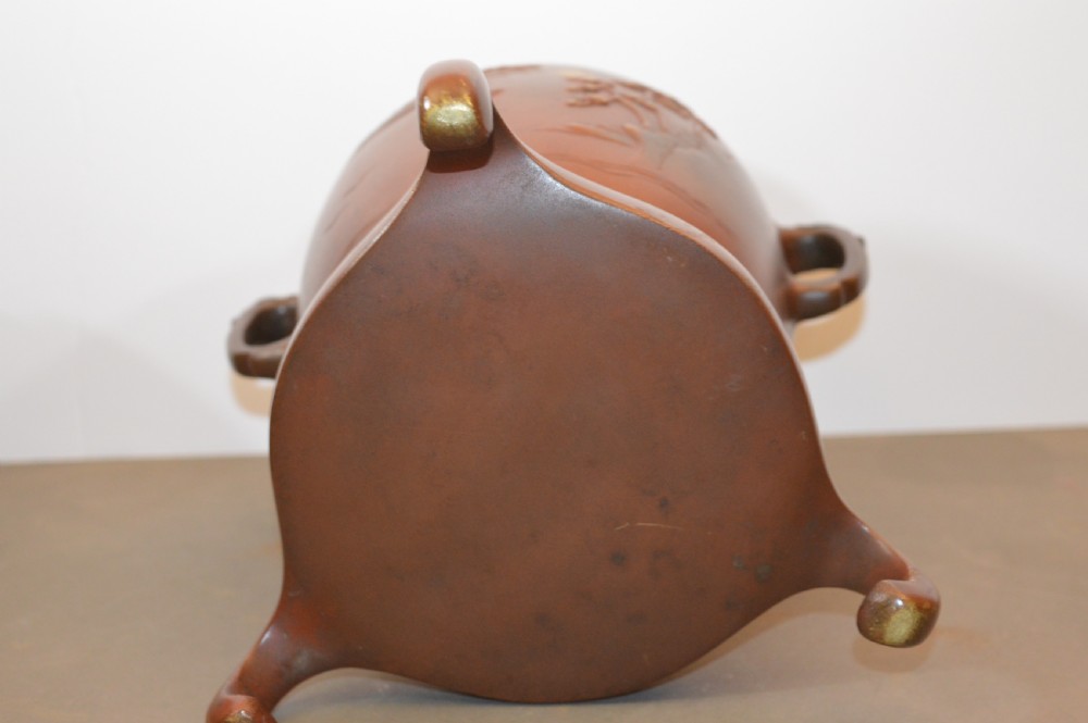 A Japanese Meiji period bronze urn featuring low relief carvings of pine trees and mountains with twin handles, resting on a matching carved bronze tripod stand. View VII