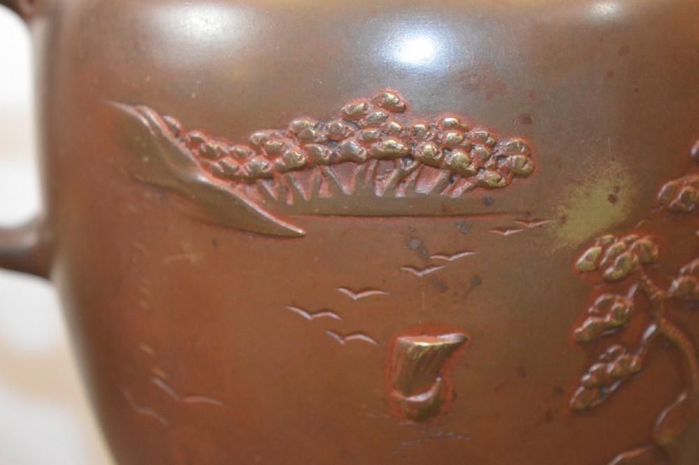 A Japanese Meiji period bronze urn featuring low relief carvings of pine trees and mountains with twin handles, resting on a matching carved bronze tripod stand. View IX