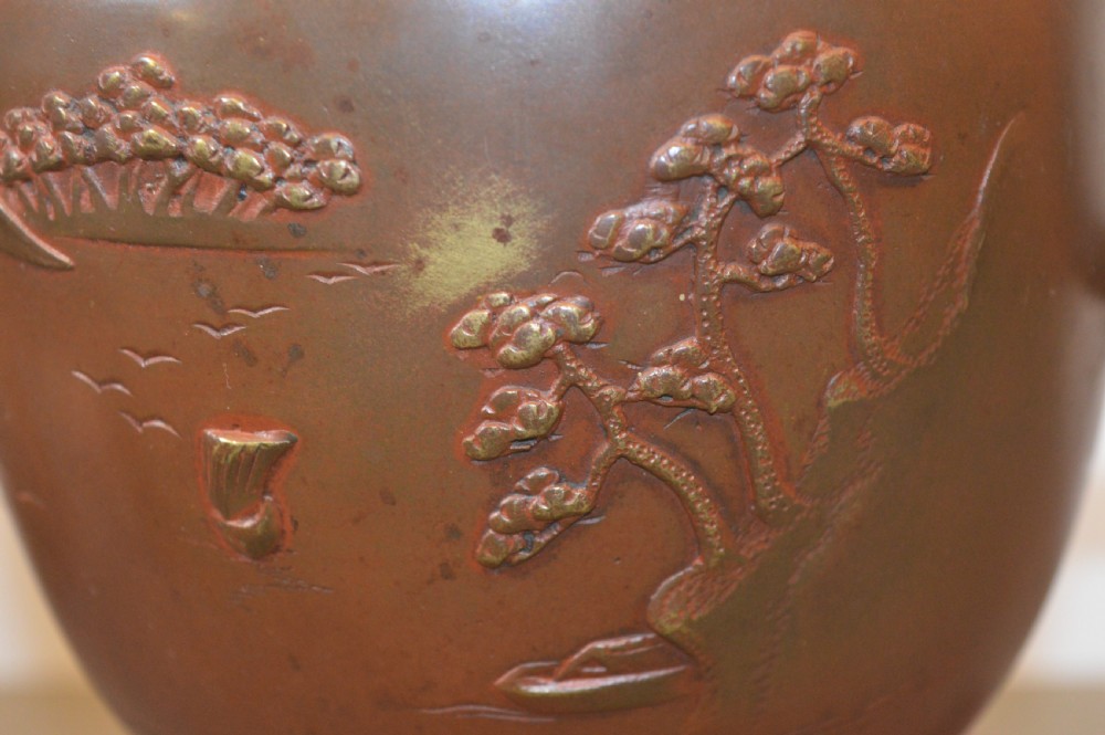 A Japanese Meiji period bronze urn featuring low relief carvings of pine trees and mountains with twin handles, resting on a matching carved bronze tripod stand. View X