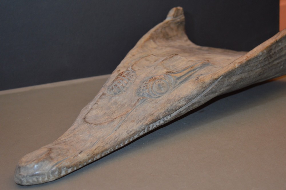 A carved wooden canoe prow shaped like a stylised crocodile head with weathered grey timber and incised geometric details around the eyes.