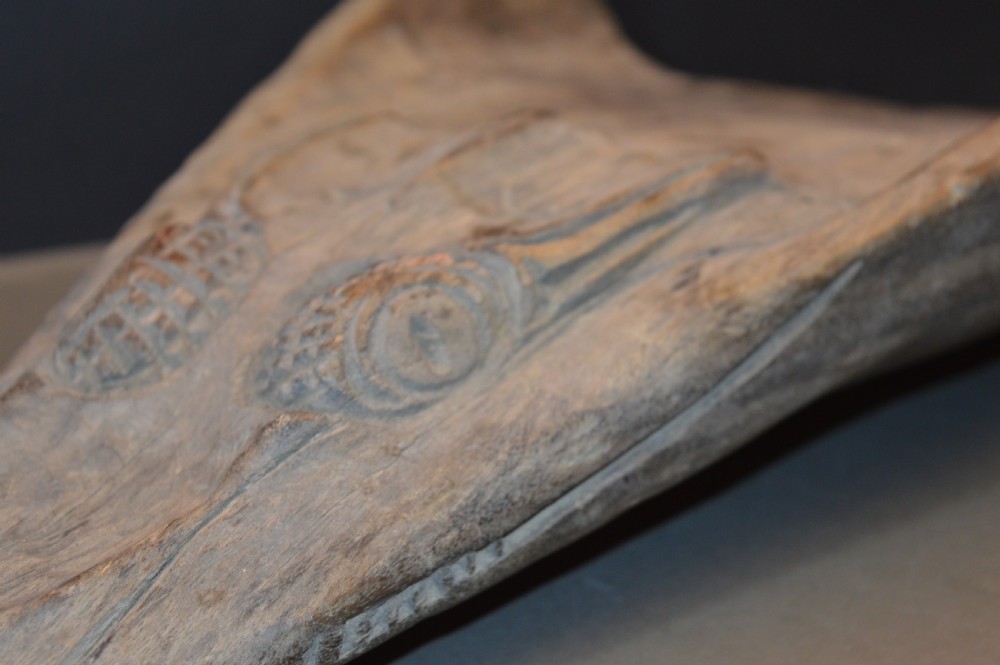 A carved wooden canoe prow shaped like a stylised crocodile head with weathered grey timber and incised geometric details around the eyes. View II
