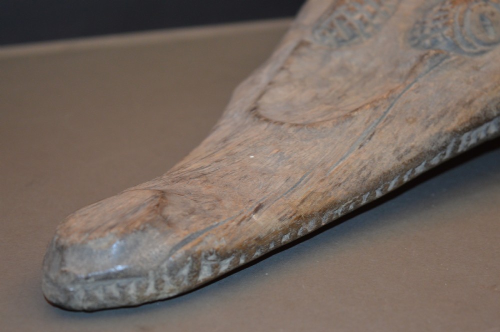 A carved wooden canoe prow shaped like a stylised crocodile head with weathered grey timber and incised geometric details around the eyes. View III