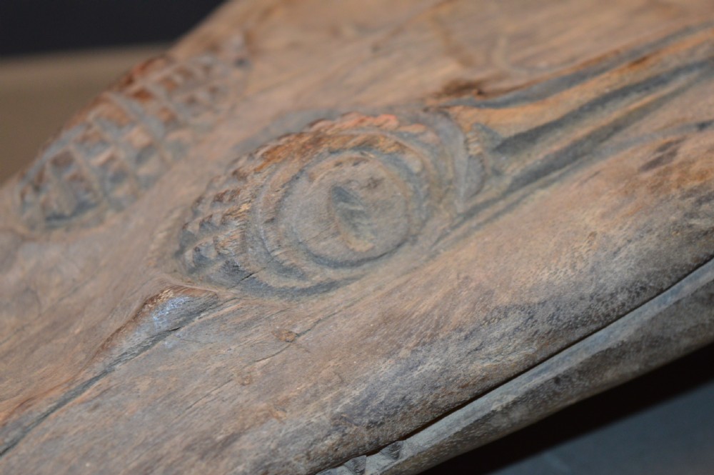 A carved wooden canoe prow shaped like a stylised crocodile head with weathered grey timber and incised geometric details around the eyes. View IV