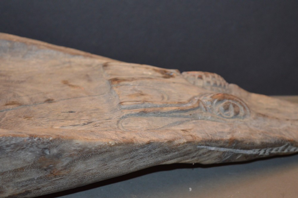 A carved wooden canoe prow shaped like a stylised crocodile head with weathered grey timber and incised geometric details around the eyes. View VI