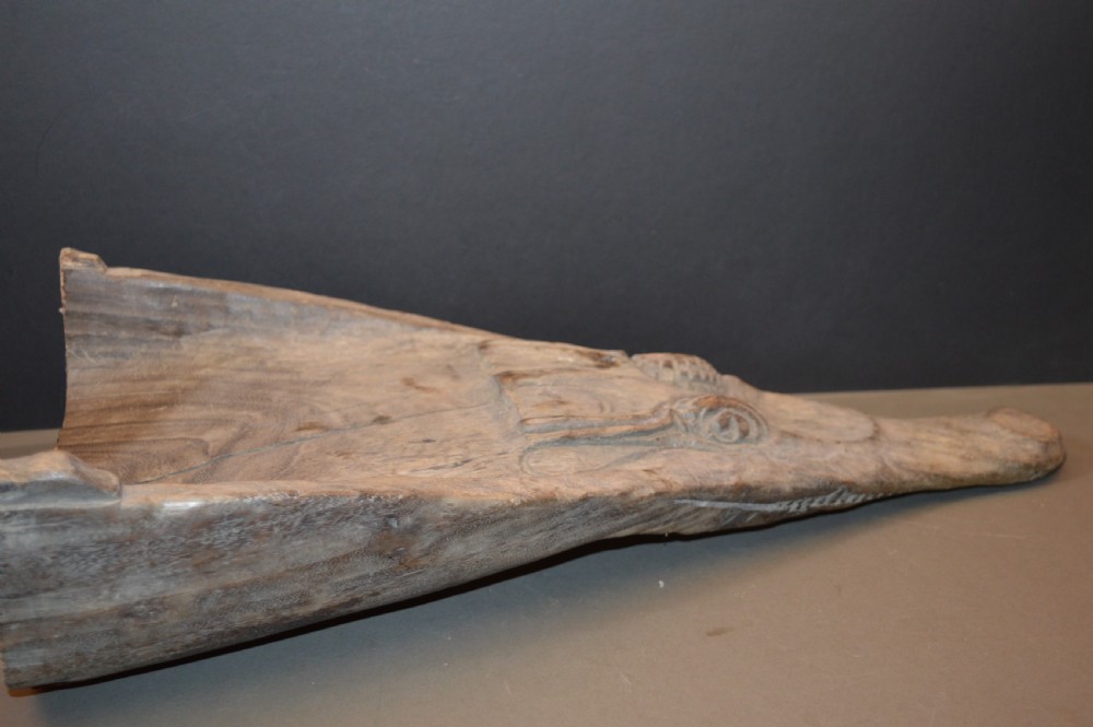 A carved wooden canoe prow shaped like a stylised crocodile head with weathered grey timber and incised geometric details around the eyes. View VII