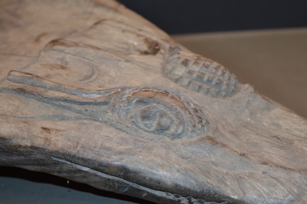 A carved wooden canoe prow shaped like a stylised crocodile head with weathered grey timber and incised geometric details around the eyes. View VIII