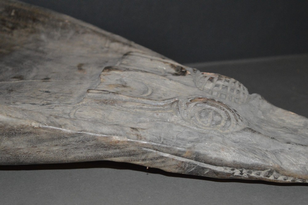 A carved wooden canoe prow shaped like a stylised crocodile head with weathered grey timber and incised geometric details around the eyes. View IX