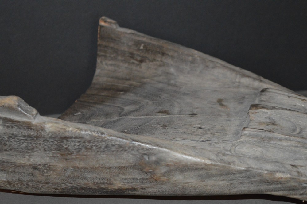 A carved wooden canoe prow shaped like a stylised crocodile head with weathered grey timber and incised geometric details around the eyes. View X