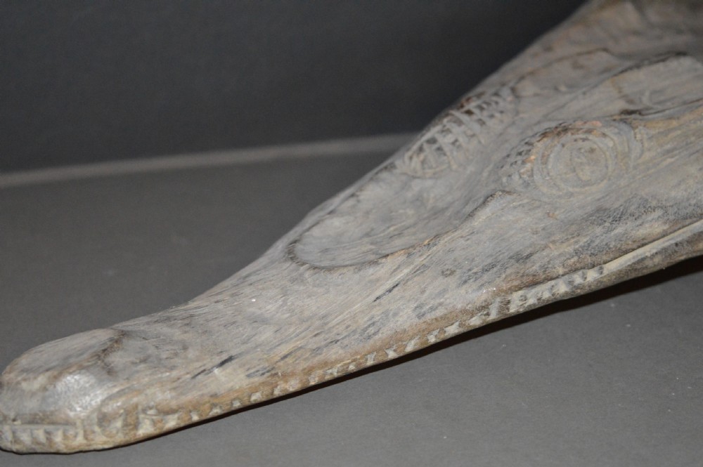 A carved wooden canoe prow shaped like a stylised crocodile head with weathered grey timber and incised geometric details around the eyes. View XII