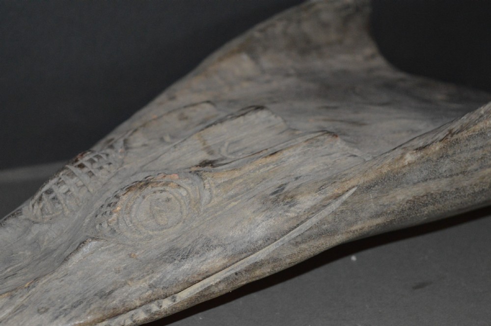 A carved wooden canoe prow shaped like a stylised crocodile head with weathered grey timber and incised geometric details around the eyes. View XIII
