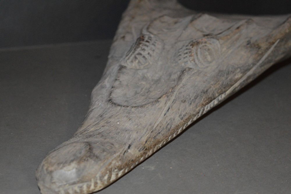 A carved wooden canoe prow shaped like a stylised crocodile head with weathered grey timber and incised geometric details around the eyes. View XXI