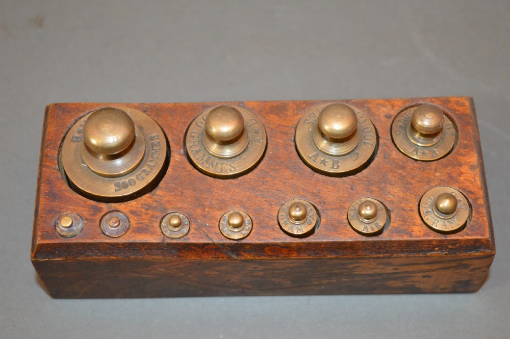 A rectangular wooden block stand fitted with a set of brass apothecary knobbed weights arranged by size. View II