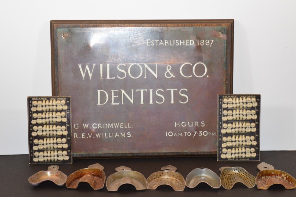 An antique framed copper dentist trade sign inscribed with business details alongside tooth sample cards and metal dental trays.
