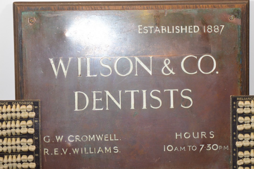 An antique framed copper dentist trade sign inscribed with business details alongside tooth sample cards and metal dental trays. View II