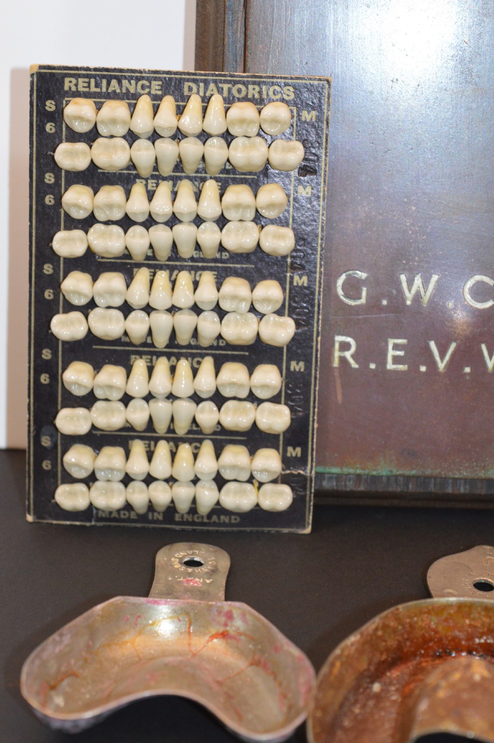 An antique framed copper dentist trade sign inscribed with business details alongside tooth sample cards and metal dental trays. View III