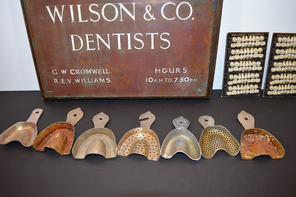 An antique framed copper dentist trade sign inscribed with business details alongside tooth sample cards and metal dental trays. View VI