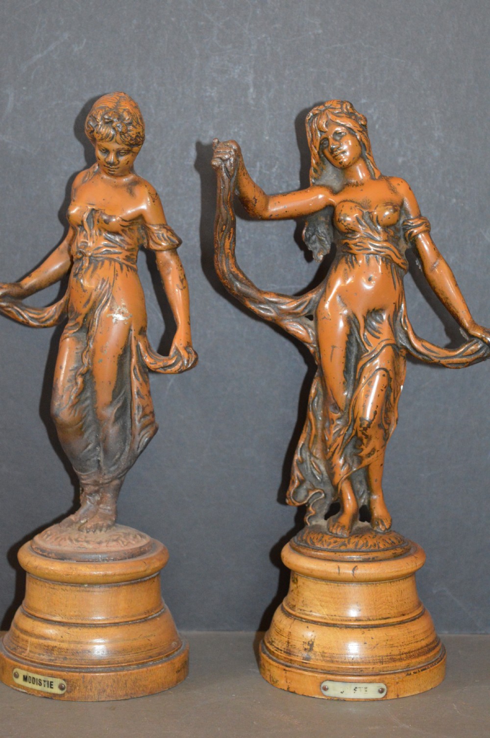 A pair of late nineteenth-century French bronzed metal allegorical female statues titled Modestie and Gaiete, mounted on circular turned wooden bases with brass nameplates.