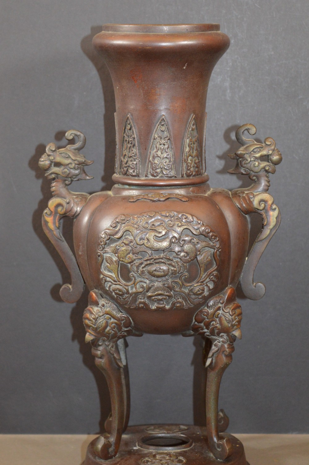 A heavy dark brown bronze Chinese incense burner featuring sculpted dragon handles, relief-carved medallions, and tall cabriole legs mounted on a round stand.