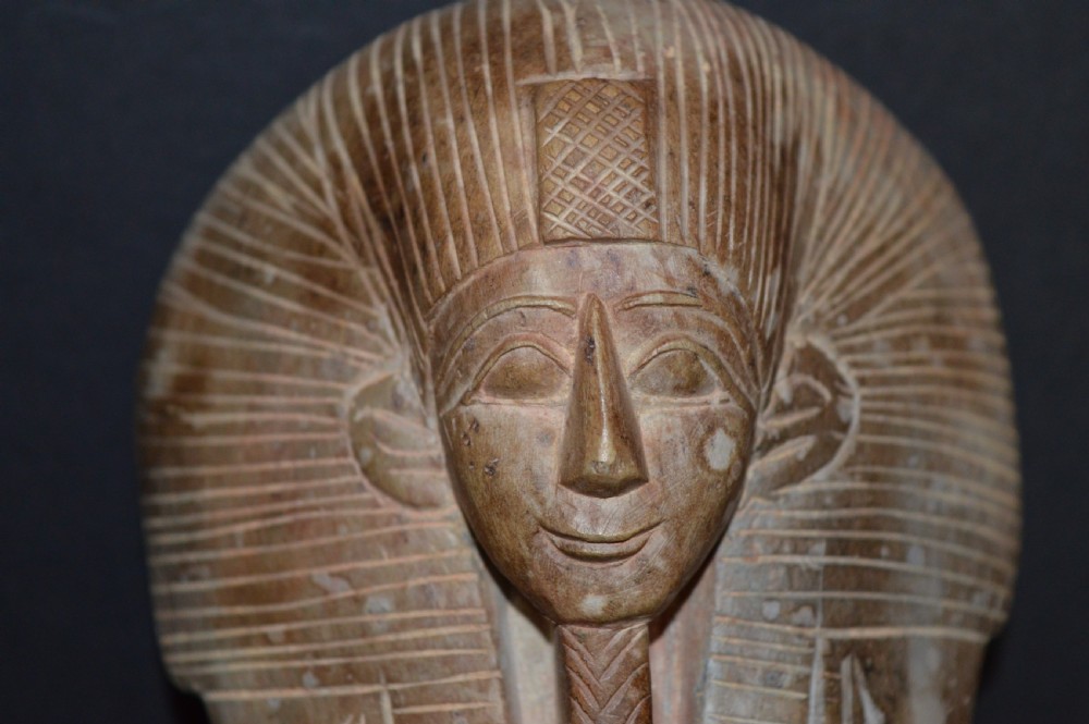 A carved beige stone model of an Egyptian sarcophagus lid featuring a pharaonic face with a nemes headdress and hieroglyphic engravings. View V