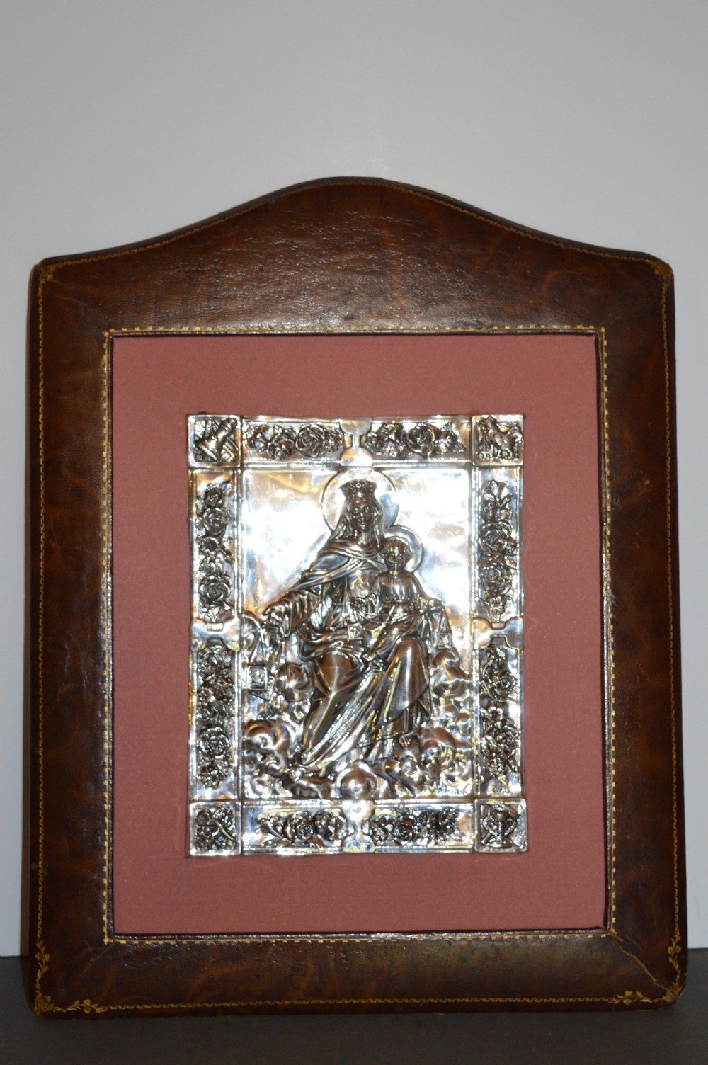 A 1920s Spanish religious icon depicting the Madonna and Child in embossed silver-toned metal, set within a brown leather frame.