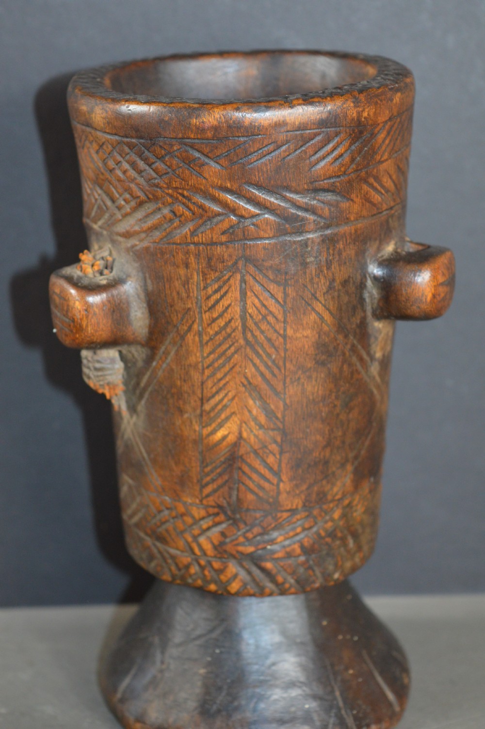 A cylindrical hand-carved dark wood vessel featuring geometric chevron patterns, side hanging lugs with fibre detail, and a flared pedestal base.