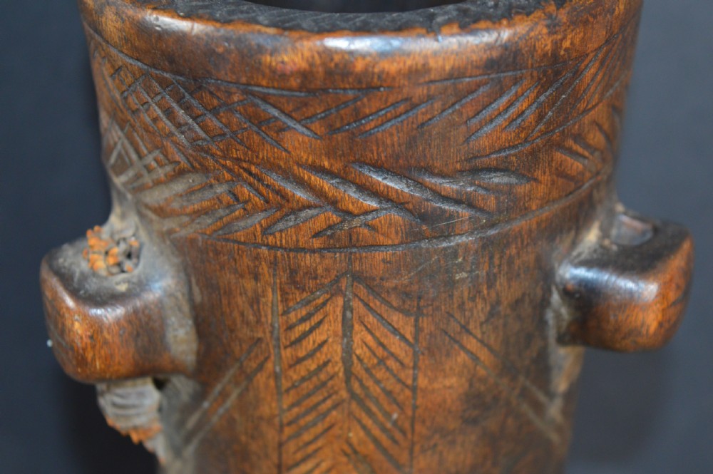 A cylindrical hand-carved dark wood vessel featuring geometric chevron patterns, side hanging lugs with fibre detail, and a flared pedestal base. View II