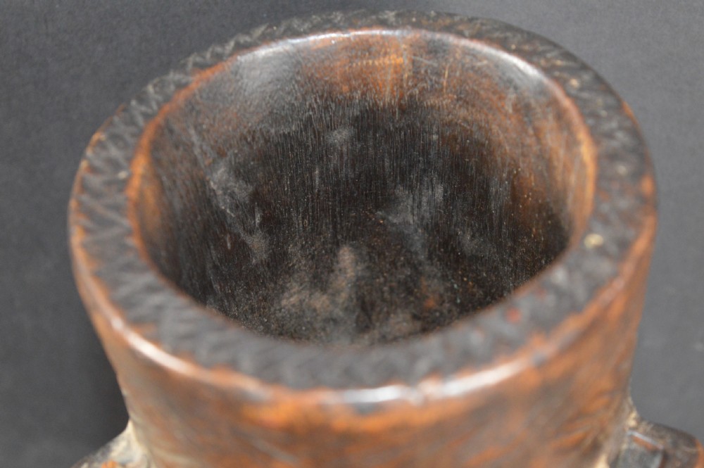 A cylindrical hand-carved dark wood vessel featuring geometric chevron patterns, side hanging lugs with fibre detail, and a flared pedestal base. View III
