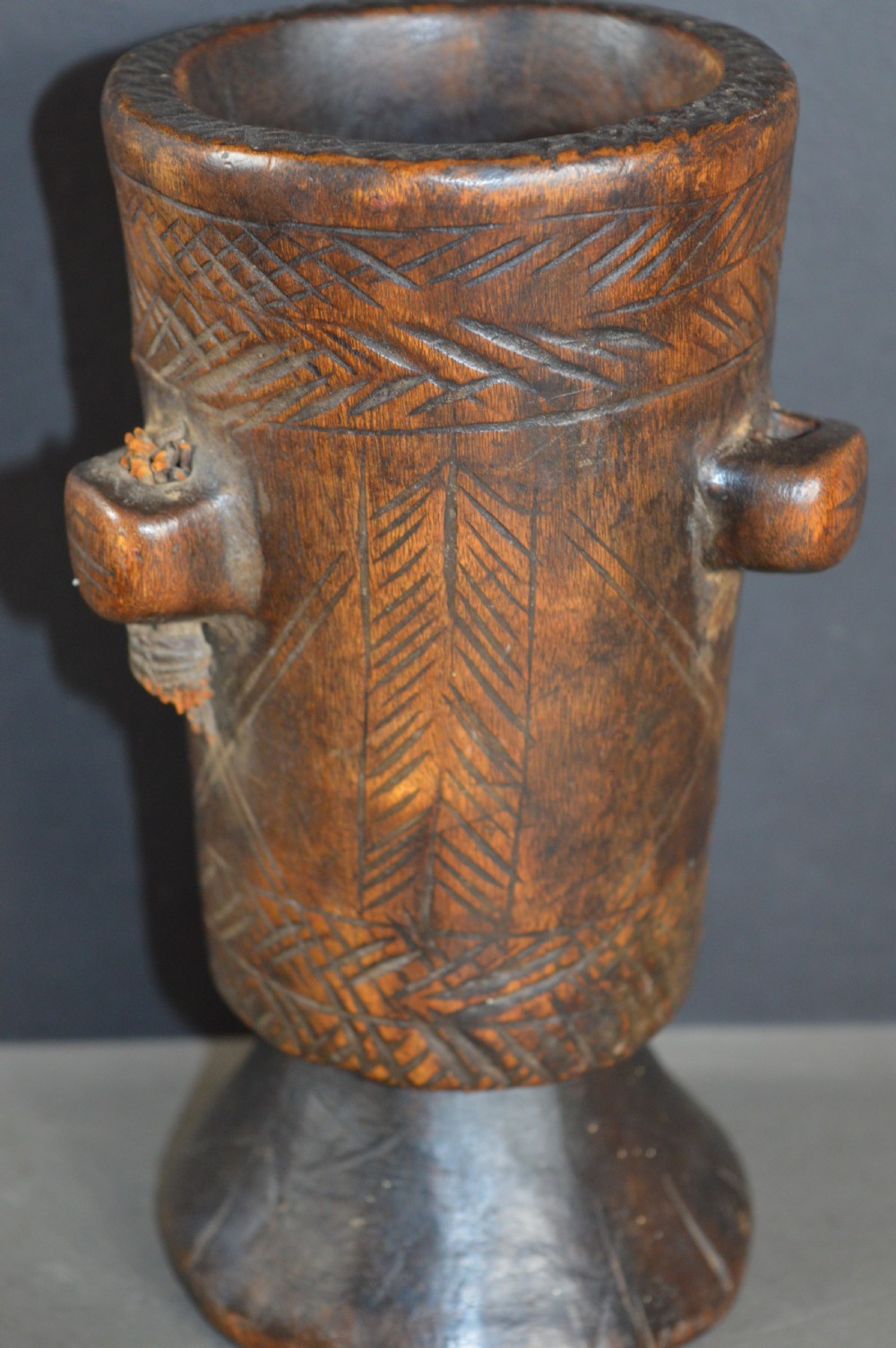 A cylindrical hand-carved dark wood vessel featuring geometric chevron patterns, side hanging lugs with fibre detail, and a flared pedestal base. View IV