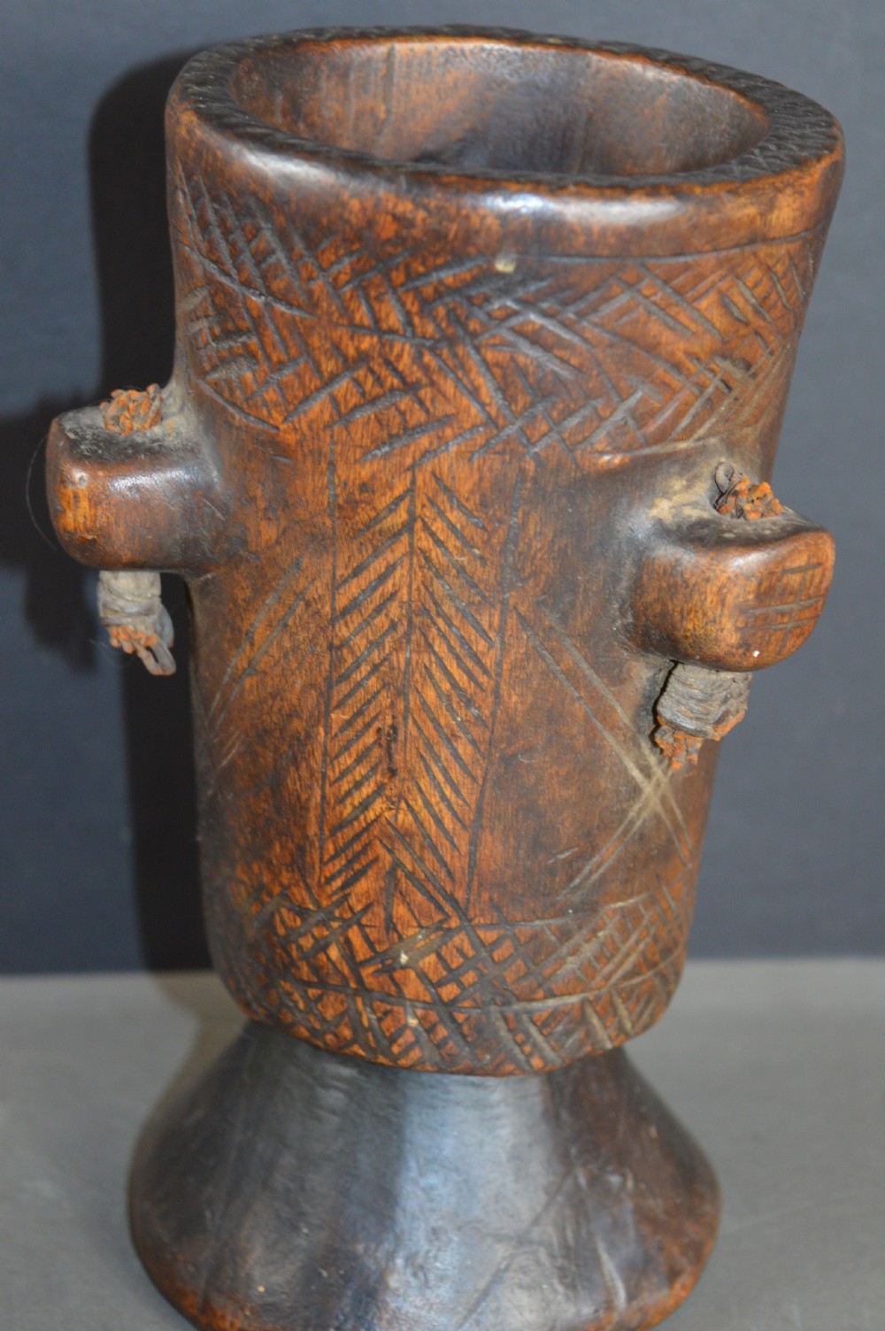 A cylindrical hand-carved dark wood vessel featuring geometric chevron patterns, side hanging lugs with fibre detail, and a flared pedestal base. View V