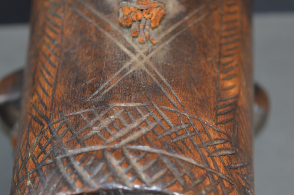 A cylindrical hand-carved dark wood vessel featuring geometric chevron patterns, side hanging lugs with fibre detail, and a flared pedestal base. View VIII