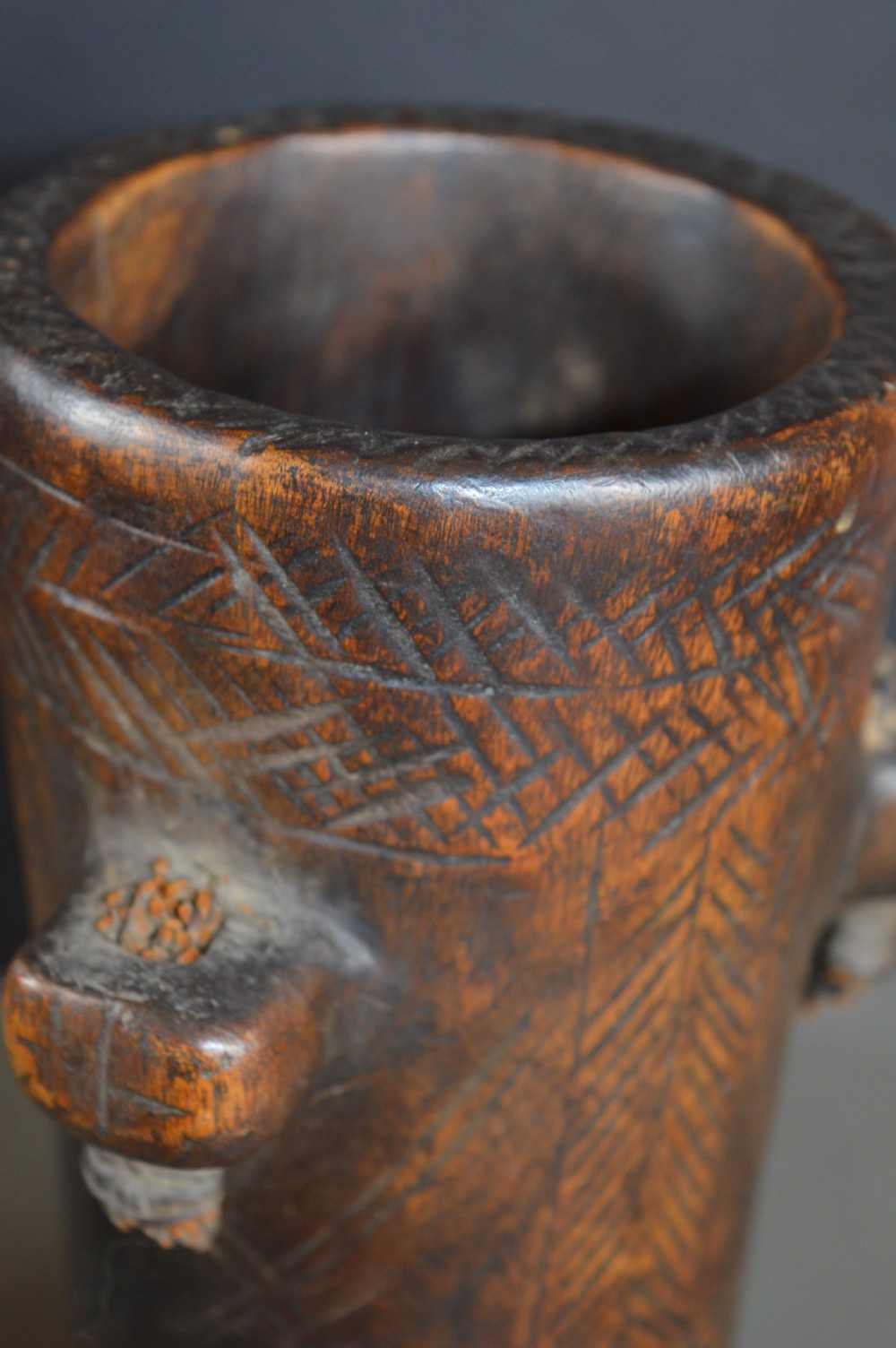 A cylindrical hand-carved dark wood vessel featuring geometric chevron patterns, side hanging lugs with fibre detail, and a flared pedestal base. View IX