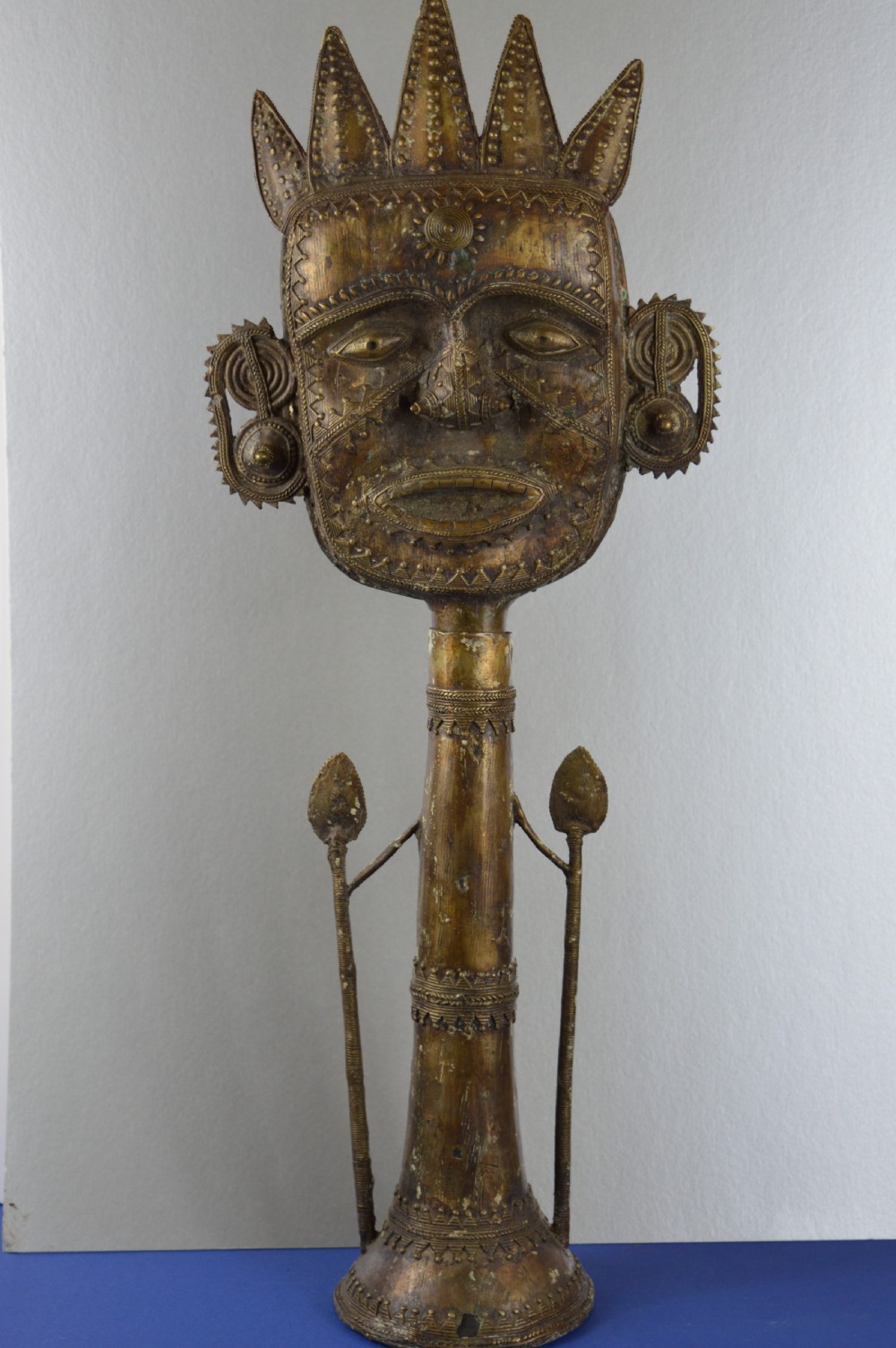 A tall, cast brass African statue featuring a stylized king figure with a spiked crown, elaborate facial details, and a tapered cylindrical base.