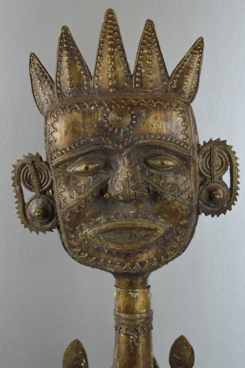 A tall, cast brass African statue featuring a stylized king figure with a spiked crown, elaborate facial details, and a tapered cylindrical base. View VI