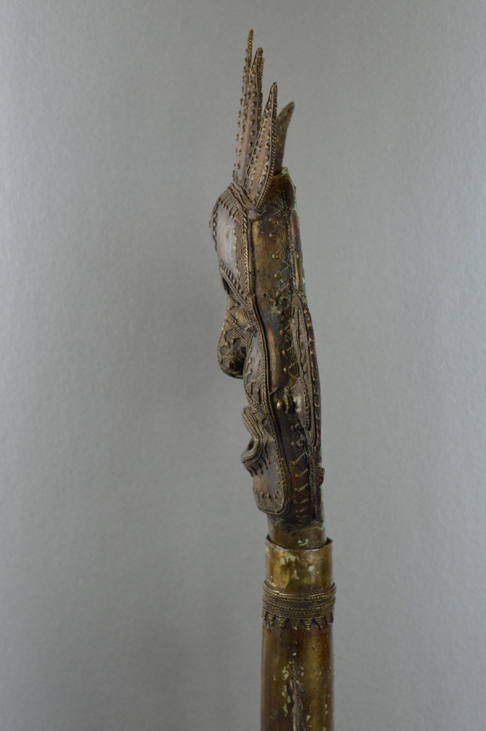 A tall, cast brass African statue featuring a stylized king figure with a spiked crown, elaborate facial details, and a tapered cylindrical base. View VII