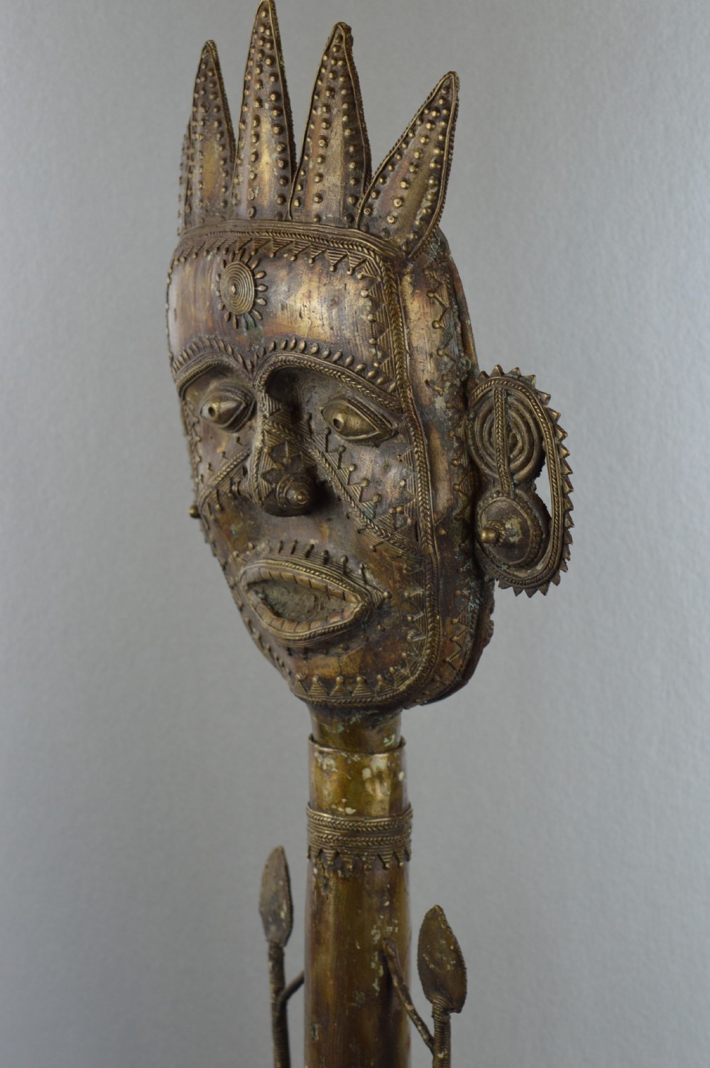 A tall, cast brass African statue featuring a stylized king figure with a spiked crown, elaborate facial details, and a tapered cylindrical base. View VIII