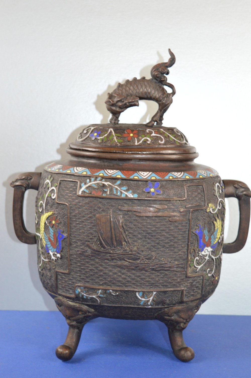 A late nineteenth-century Japanese bronze tripod censer featuring polychrome cloisonne enamel work, relief panels depicting a sailboat, loop handles, and a cover topped with a beast finial.