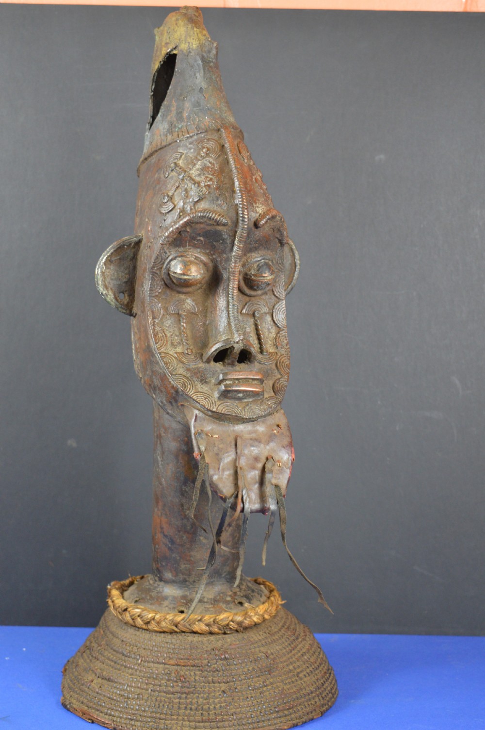A tall dark bronze Yoruba head displaying facial scarification, a high conical helmet, leather beard attachments, and a woven rope base.