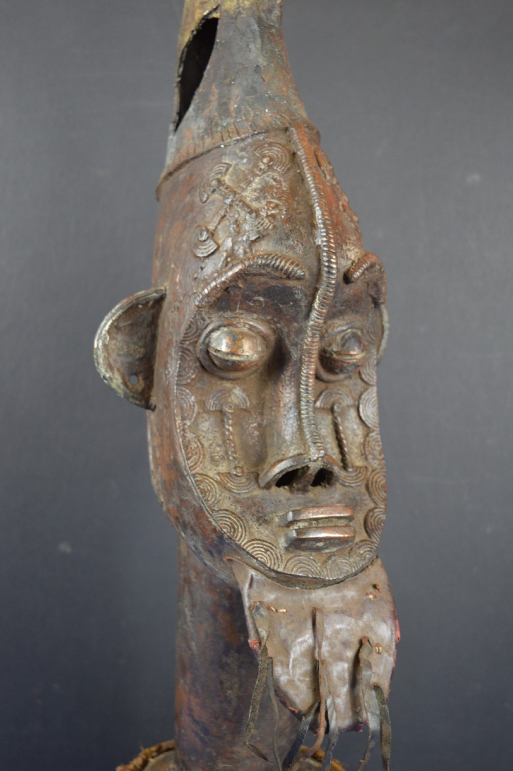 A tall dark bronze Yoruba head displaying facial scarification, a high conical helmet, leather beard attachments, and a woven rope base. View II