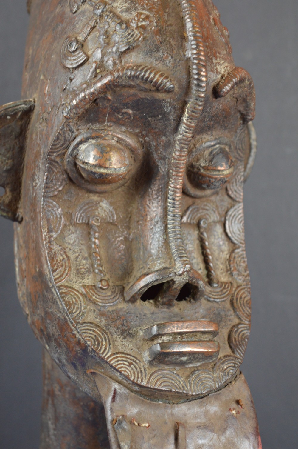 A tall dark bronze Yoruba head displaying facial scarification, a high conical helmet, leather beard attachments, and a woven rope base. View III