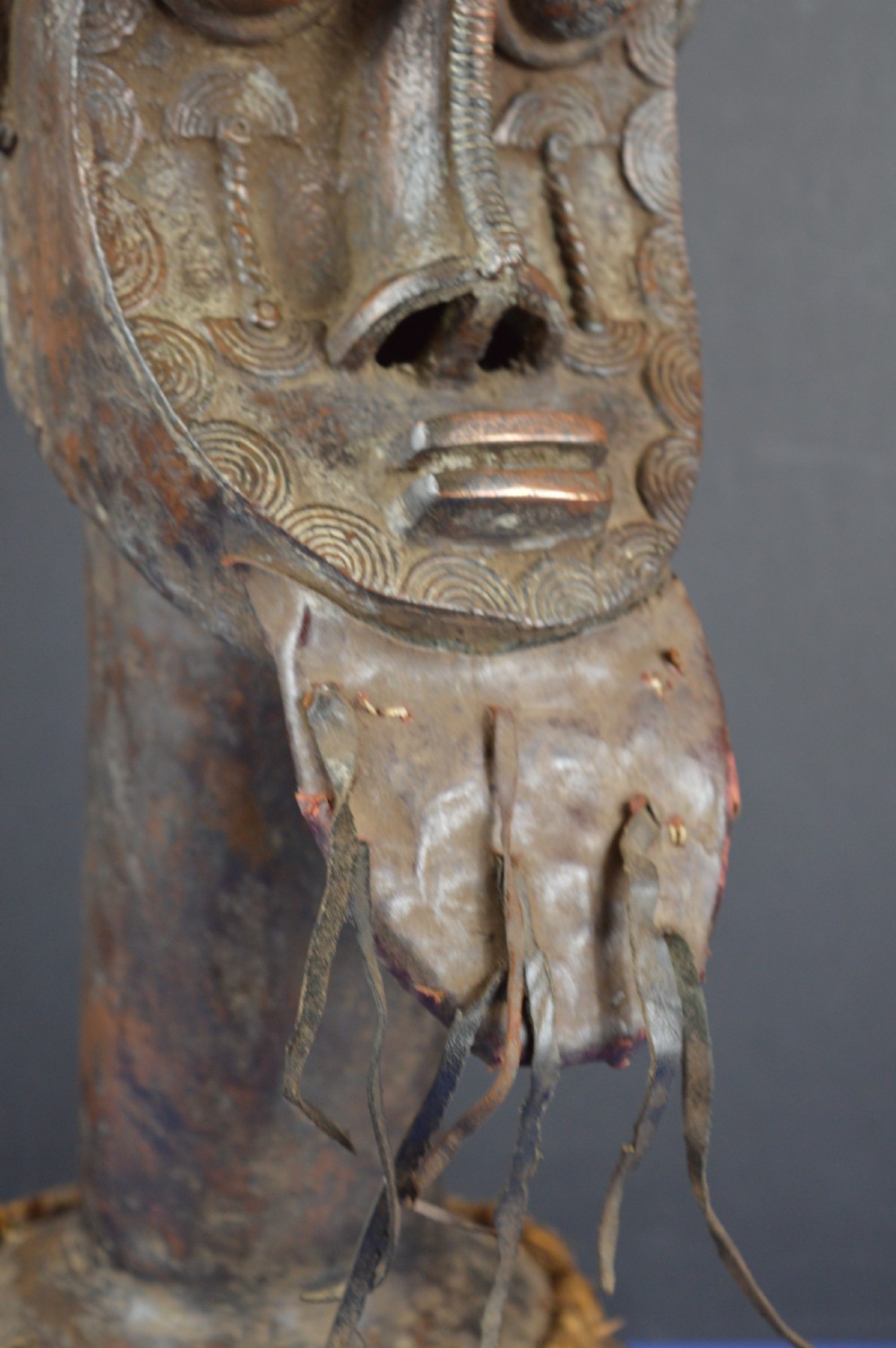 A tall dark bronze Yoruba head displaying facial scarification, a high conical helmet, leather beard attachments, and a woven rope base. View V
