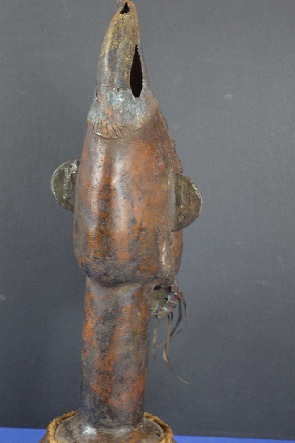 A tall dark bronze Yoruba head displaying facial scarification, a high conical helmet, leather beard attachments, and a woven rope base. View VII