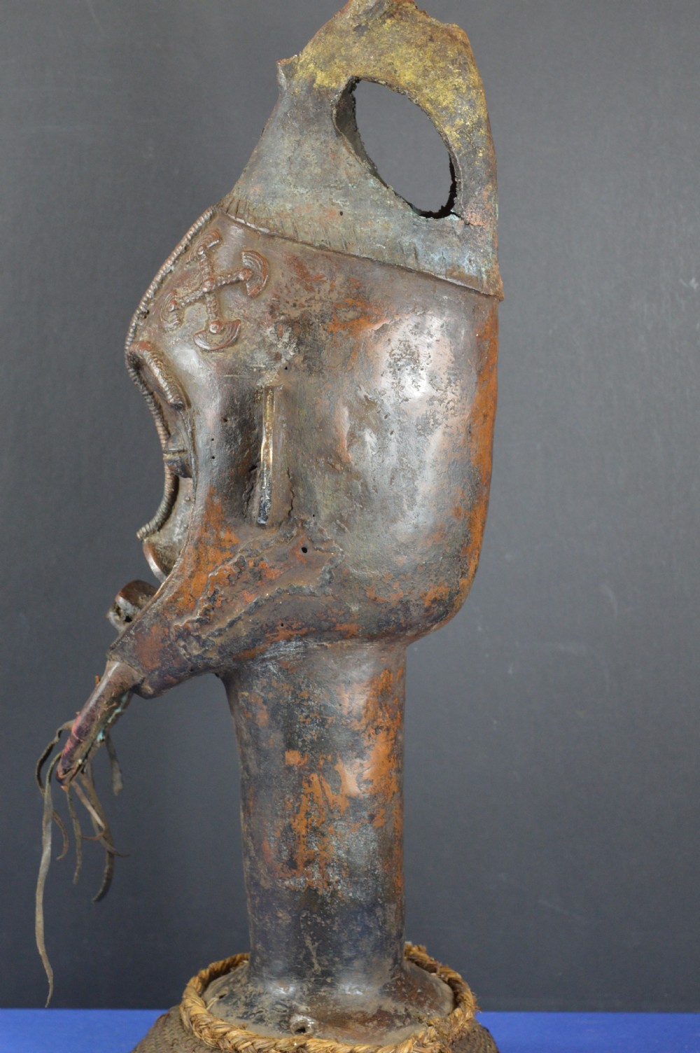 A tall dark bronze Yoruba head displaying facial scarification, a high conical helmet, leather beard attachments, and a woven rope base. View VIII