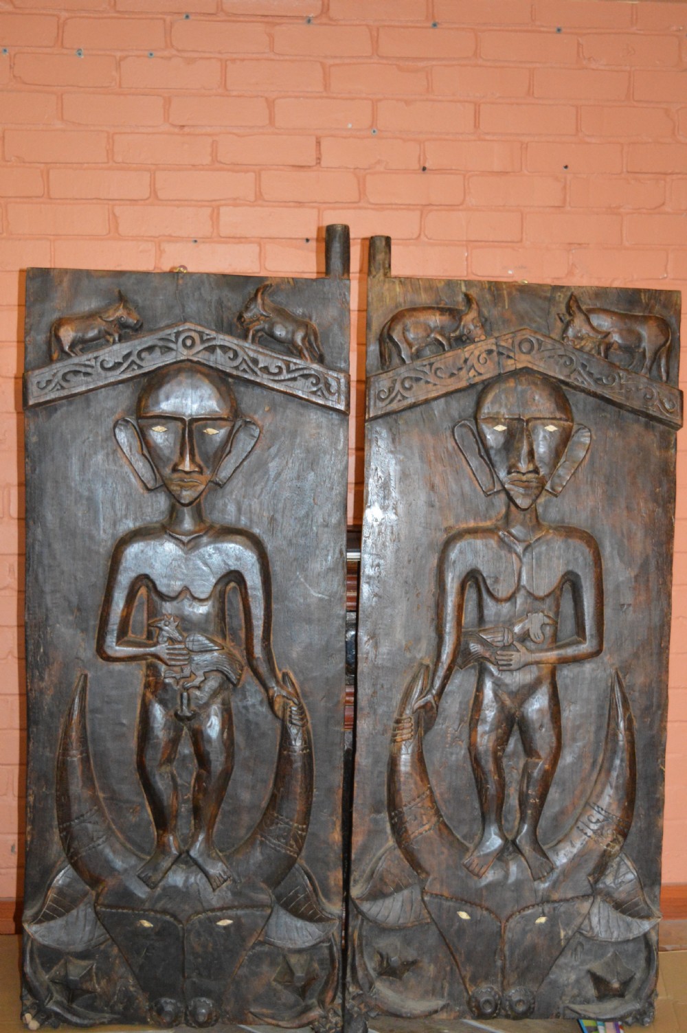 Pair of dark hand-carved wooden doors featuring high-relief human figures holding roosters above water buffalo heads with bone inlaid eyes.