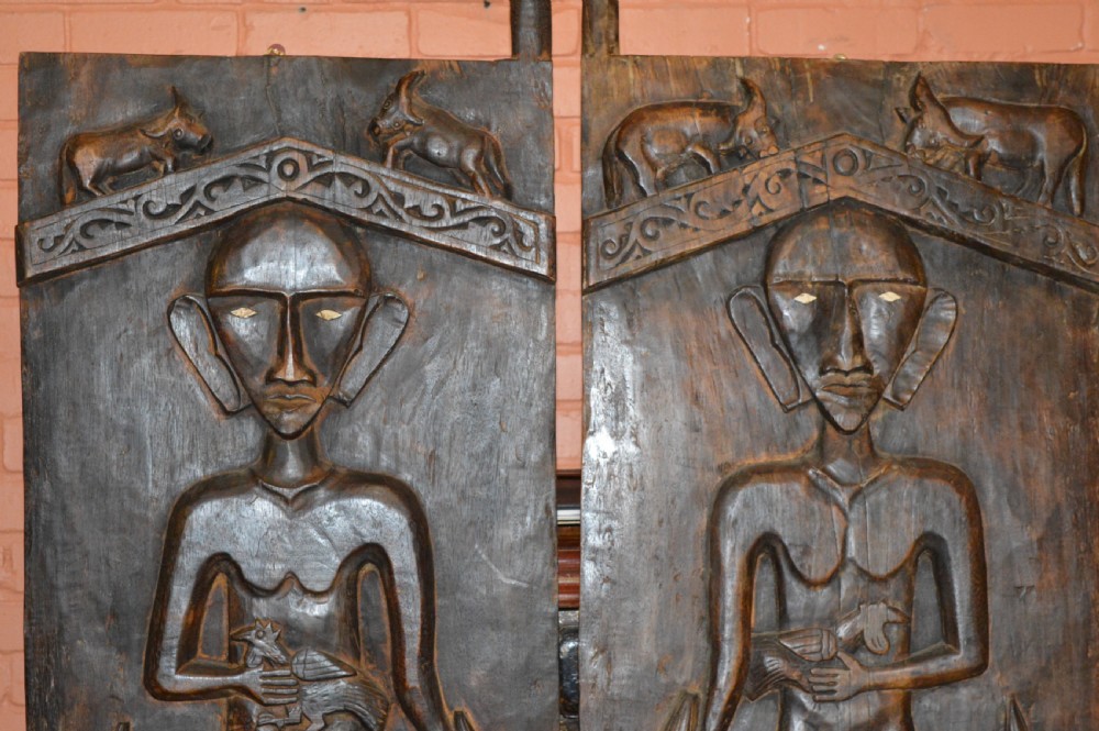 Pair of dark hand-carved wooden doors featuring high-relief human figures holding roosters above water buffalo heads with bone inlaid eyes. View II