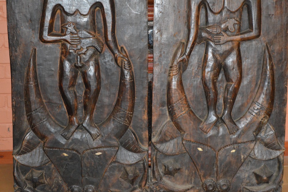 Pair of dark hand-carved wooden doors featuring high-relief human figures holding roosters above water buffalo heads with bone inlaid eyes. View III