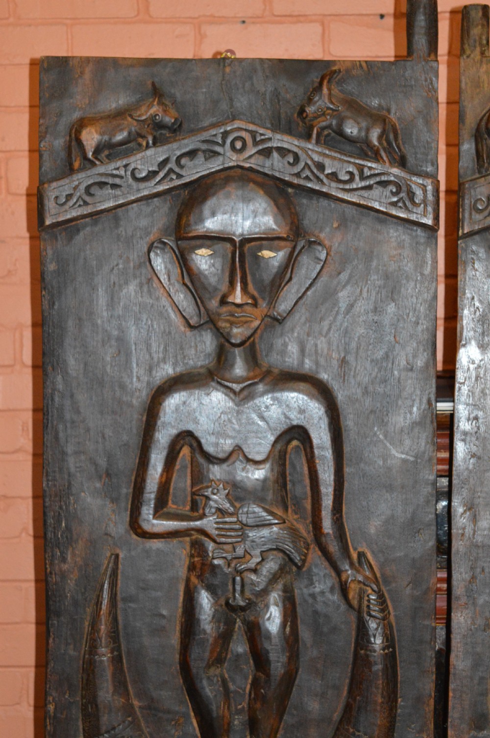 Pair of dark hand-carved wooden doors featuring high-relief human figures holding roosters above water buffalo heads with bone inlaid eyes. View IV