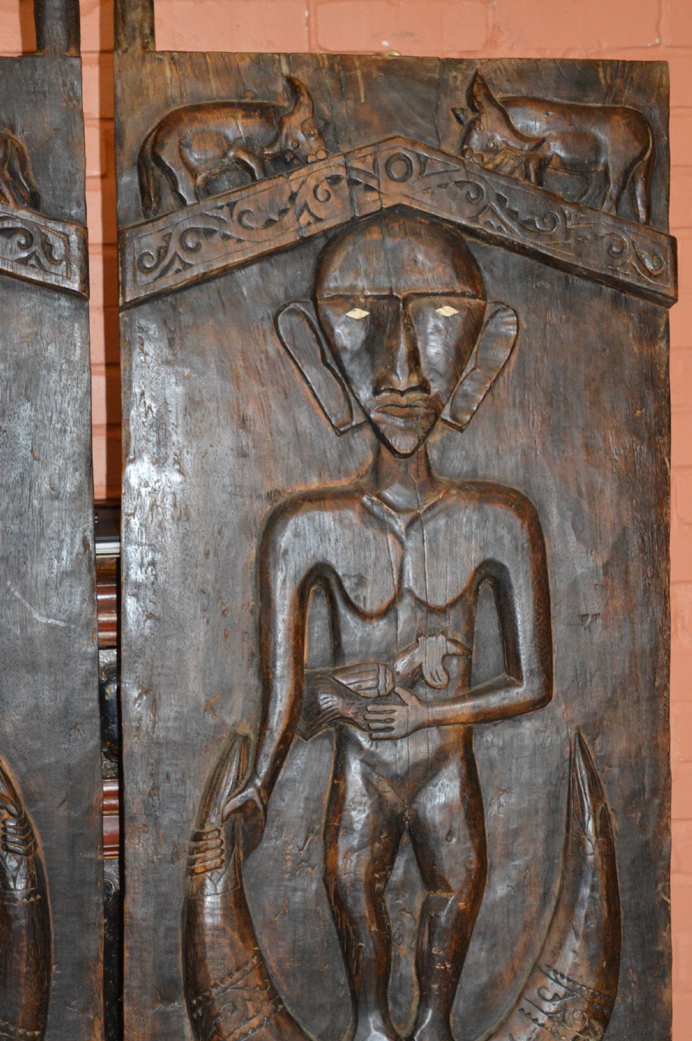 Pair of dark hand-carved wooden doors featuring high-relief human figures holding roosters above water buffalo heads with bone inlaid eyes. View V