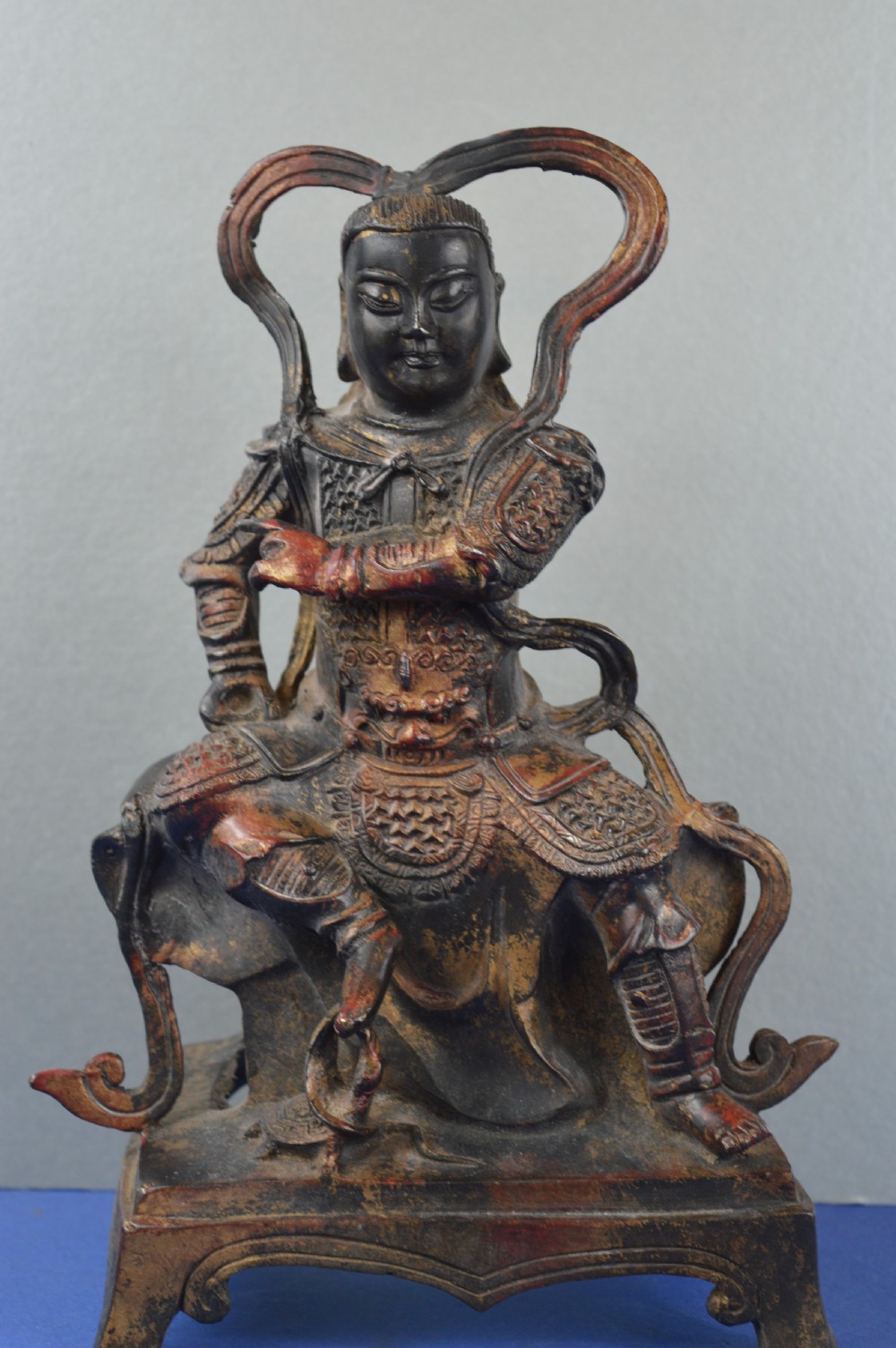 A late nineteenth-century Chinese dark bronze sculpture depicting a seated warrior in detailed armour with polychrome red accents and a decorative halo motif.