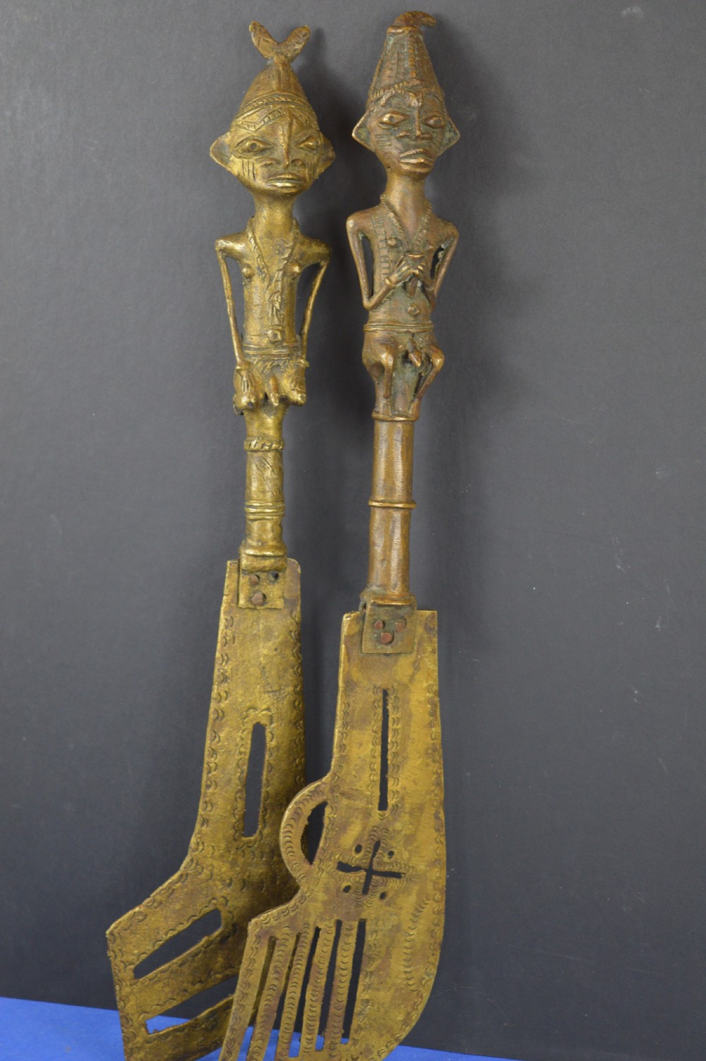 A pair of cast bronze Yoruba ceremonial staffs featuring sculpted human figures as finials above flat, patterned brass blades.