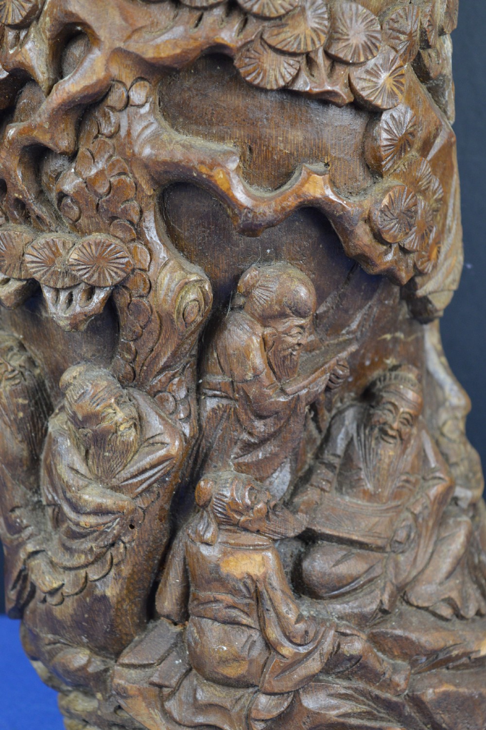 A hand-carved cylindrical bamboo brush pot featuring figures and pine trees carved in high relief against a warm brown wooden surface. View XI